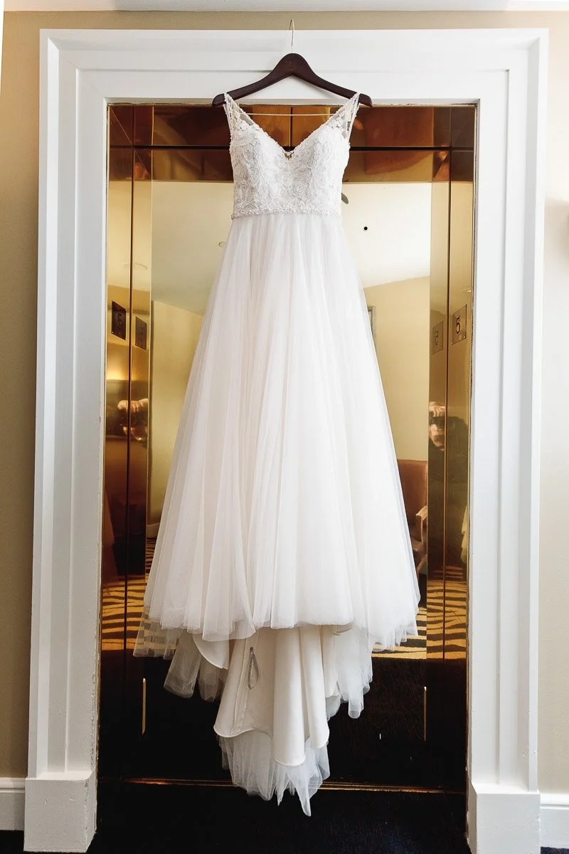 Elegant white wedding dress with lace bodice and flowing tulle skirt hanging on a wooden hanger in a warmly lit room with gold-toned accents.
