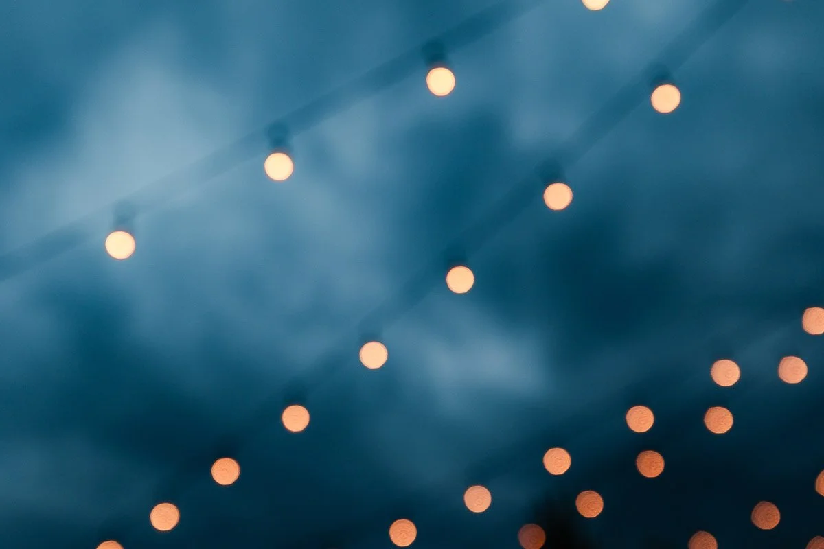 String lights softly blur against a moody, cloudy evening sky. The warm glow of the lights contrasts with the cool, dark atmosphere, creating a serene, dreamy ambiance.