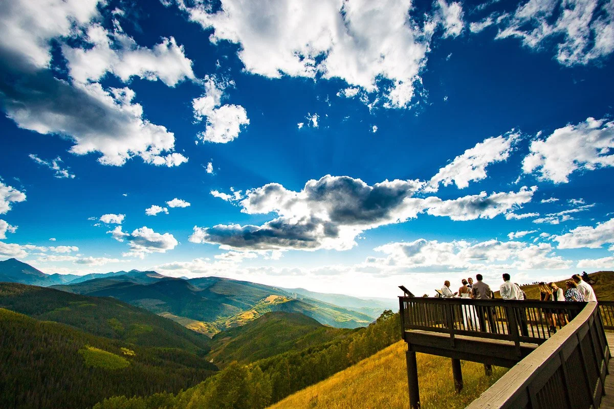 A scenic view from a lookout point, showcasing mountains and valleys under a blue sky filled with clouds, with several people standing for a Vail wedding ceremony on a wooden deck.