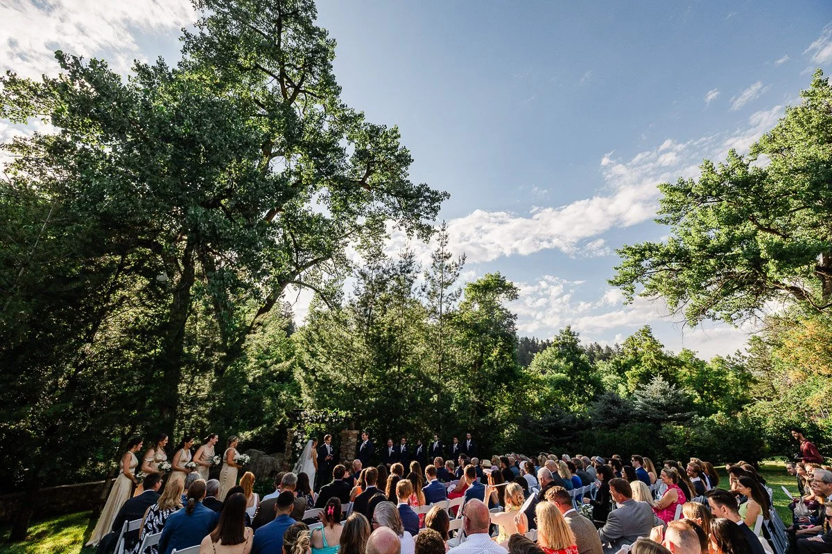 Outdoor wedding ceremony in a lush garden setting under a clear blue sky. Guests are seated facing the bridal party, surrounded by tall, green trees.