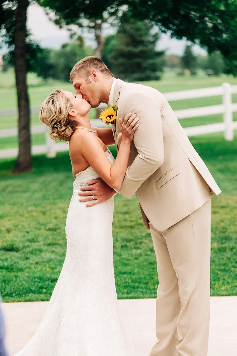 A bride and groom kiss outdoors. The bride wears a white dress and holds a sunflower, while the groom is in a light beige suit. Background shows green grass and trees.