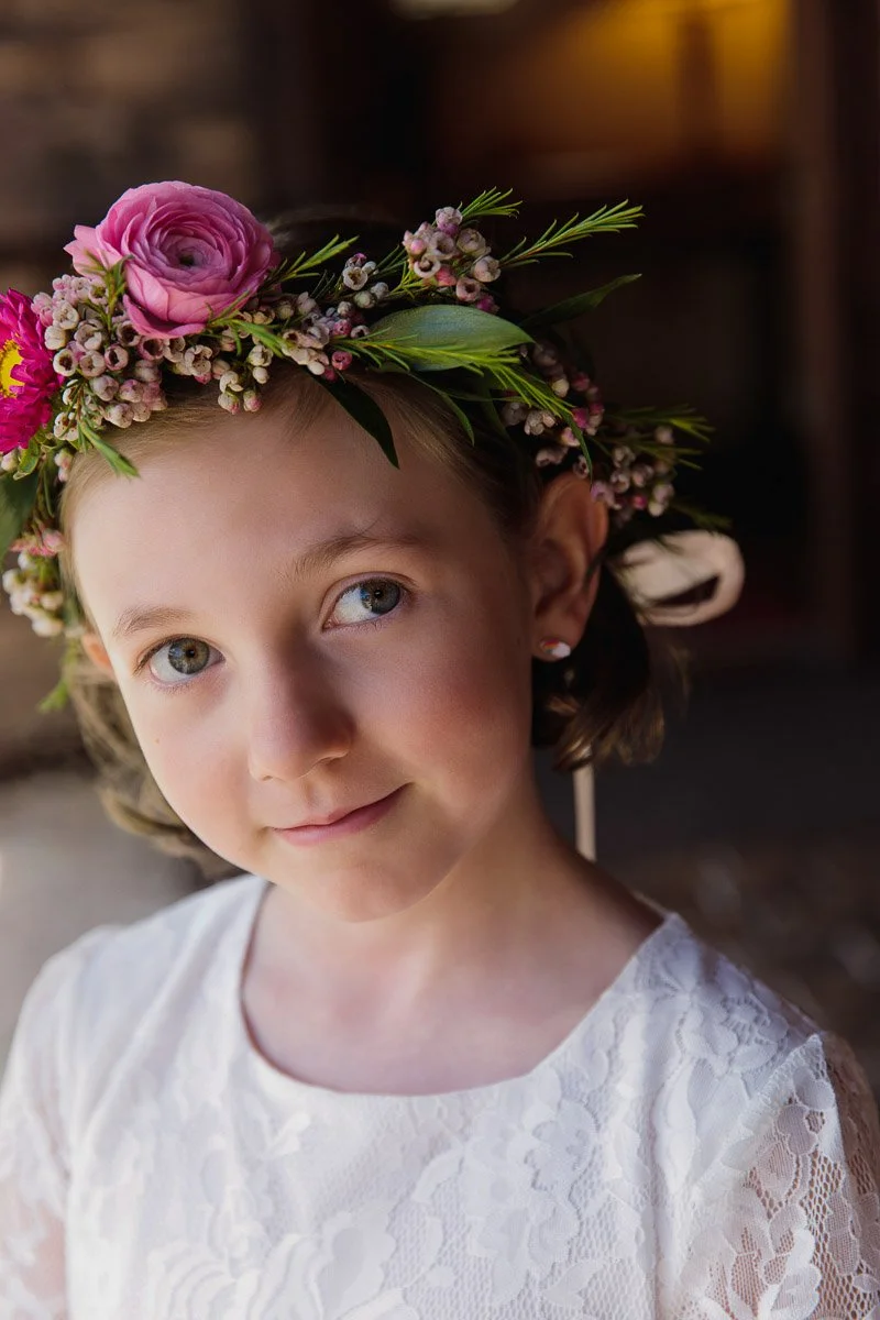 Young girl in a white lace dress, wearing a flower crown with pink blooms and greenery. She smiles softly, conveying a gentle and serene mood.