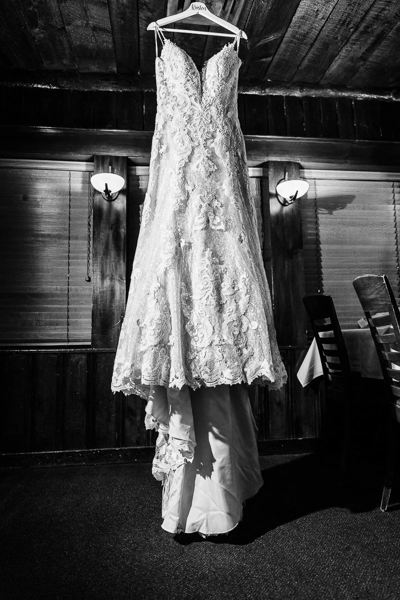 A lace wedding dress hangs elegantly on a wooden hanger in a dimly-lit rustic room with wooden walls and two wall lamps, evoking a vintage feel.