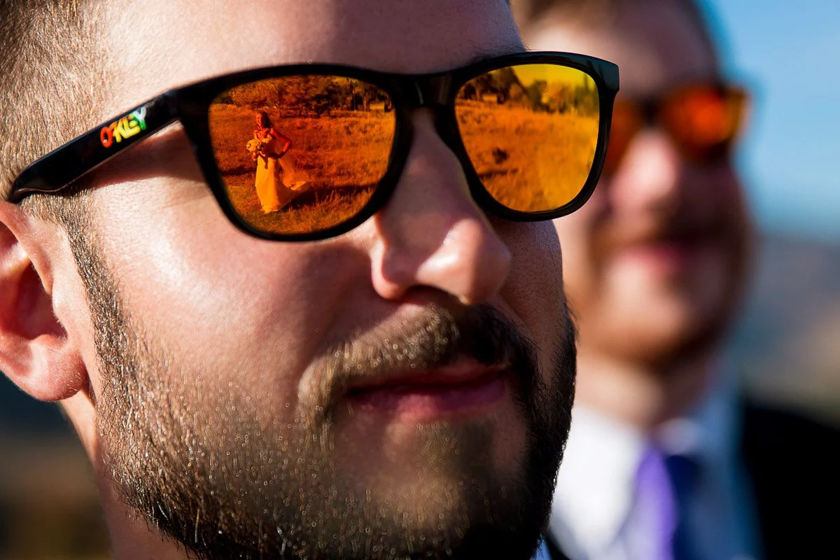 Close-up of a man wearing sunglasses with orange-tinted lenses. A bride is reflected in the lenses, set in a sunny outdoor landscape, creating a joyful tone.