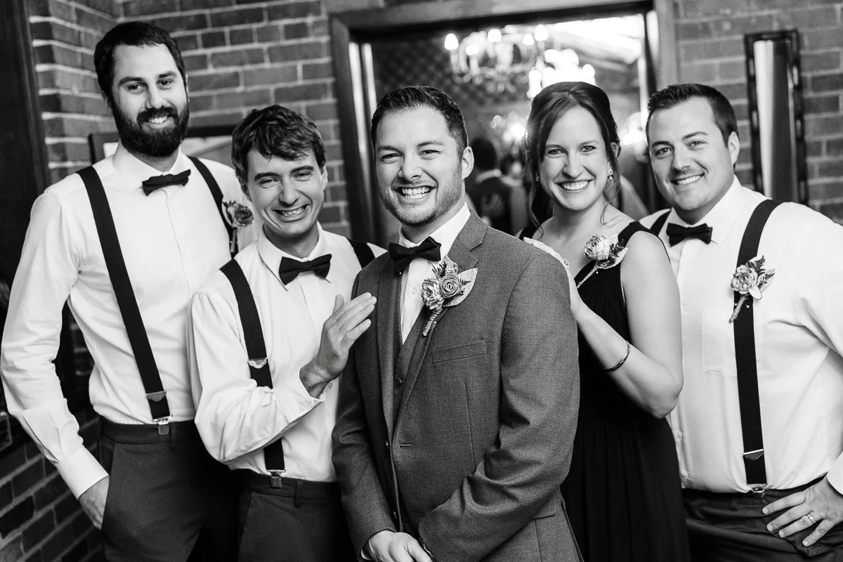 A group of five people, dressed formally with boutonnieres, smile warmly at the camera. They stand in front of a brick wall, conveying a celebratory mood.