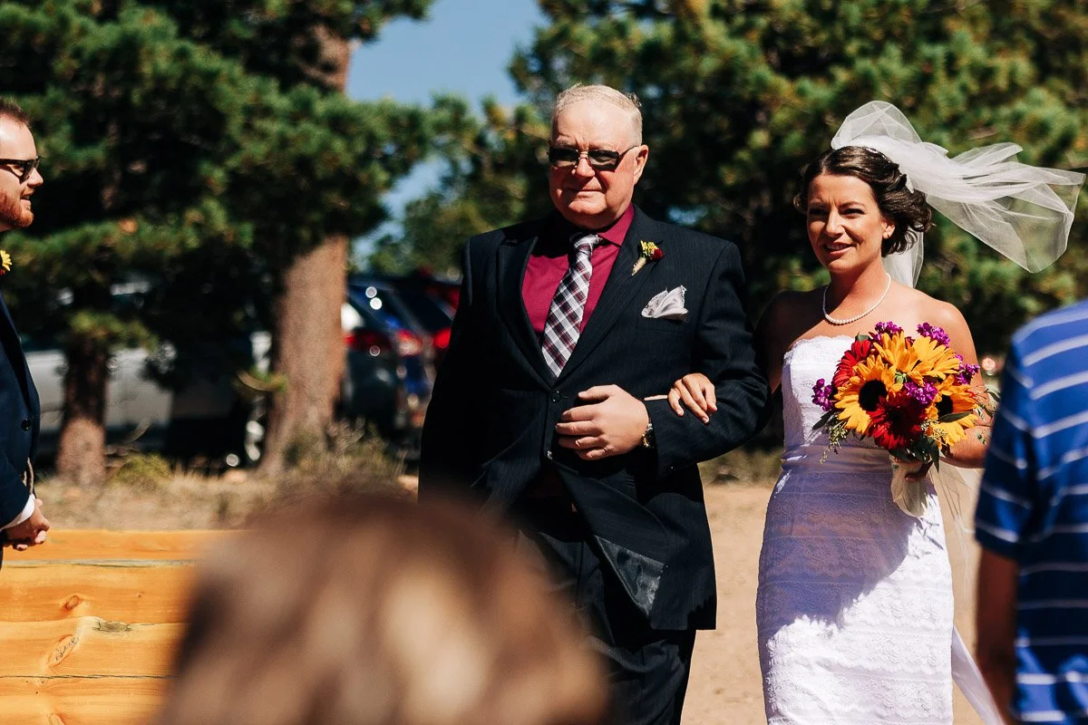 A bride in a white dress and veil walks arm-in-arm with a man in a suit down an outdoor aisle. She holds a bouquet of vibrant flowers.