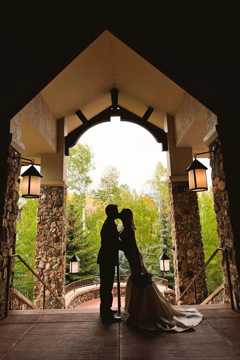 Silhouetted couple in wedding attire kissing under a stone archway, with lush green trees and soft lanterns creating a romantic, serene backdrop captured by Beaver Creek wedding photographer tomKphoto