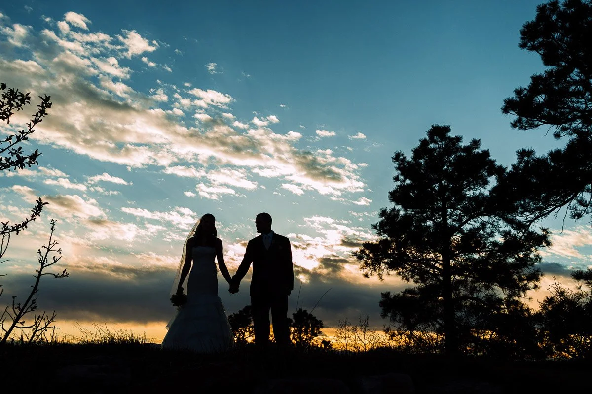 Silhouetted couple holding hands at sunset, framed by trees and a vibrant sky with clouds, creates a serene and romantic atmosphere.