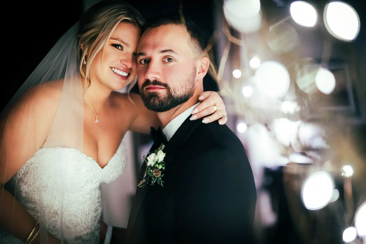 A joyous couple on their wedding day, the bride in a lace gown and veil leans affectionately on the groom in a black suit, with soft lights in the background captured by Beaver Creek wedding photographer tomKphoto