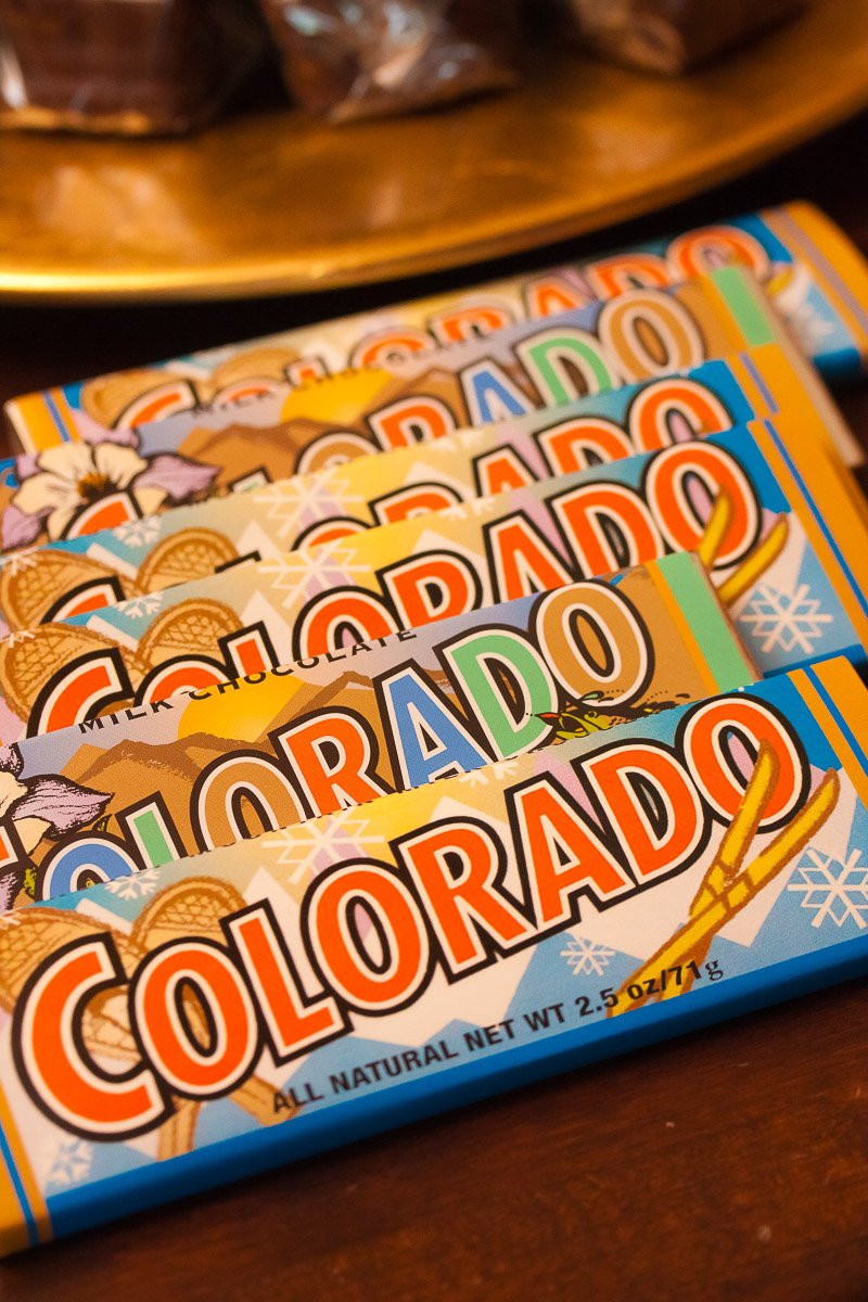 Colorful chocolate bars labeled "Colorado" are stacked in a fan shape. The design features mountains, snowflakes, and skis, evoking a winter theme.
