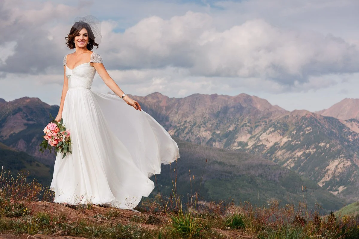 A smiling bride in a flowing white gown holds pink roses, standing on a mountain with a cloudy sky and rugged Gore Ranch peaks in the background during a Four Seasons Resort wedding in Vail, Colorado