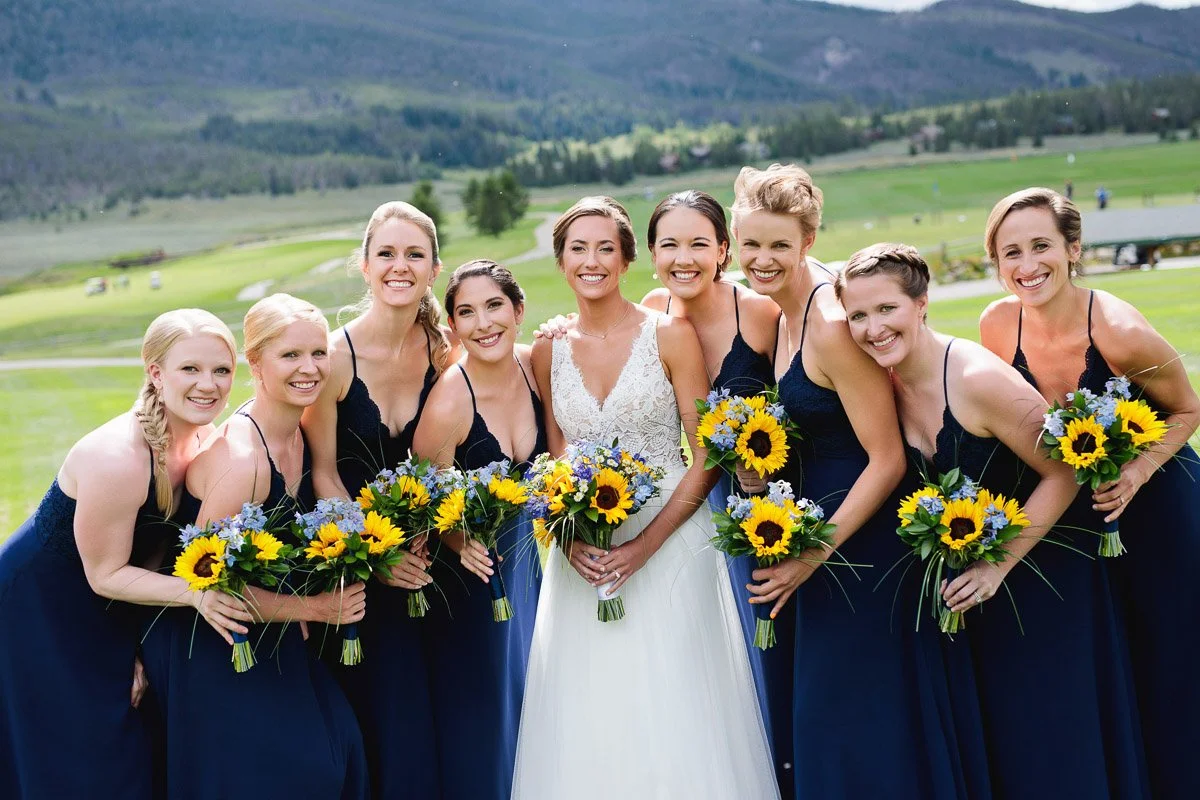 A joyful bride stands with eight bridesmaids in navy dresses on a lush green field. They hold bouquets of sunflowers, all smiling warmly. Mountains form the backdrop.