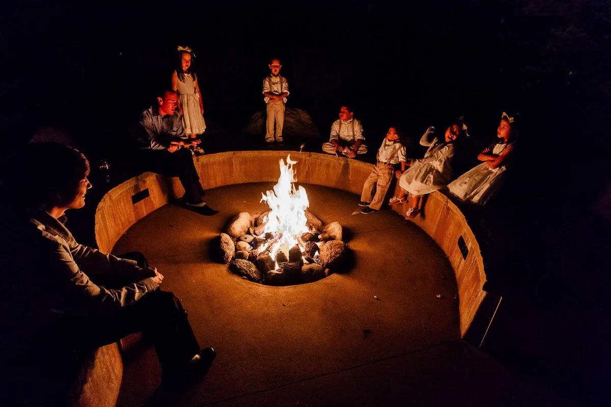 A group of people, including children and adults, are seated around a glowing campfire at night in a circular seating area, creating a cozy, warm atmosphere.