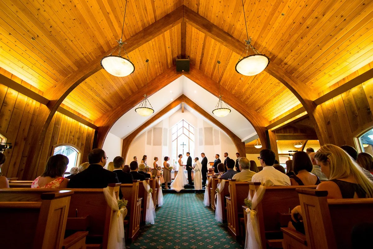 A wedding ceremony in a warmly lit Interfaith Chapel. The bride and groom stand at the altar with the bridal party, surrounded by seated guests.