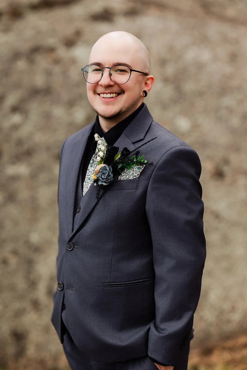 A smiling person in a gray suit with a floral tie and boutonniere stands in front of a textured rock background, conveying a cheerful tone.