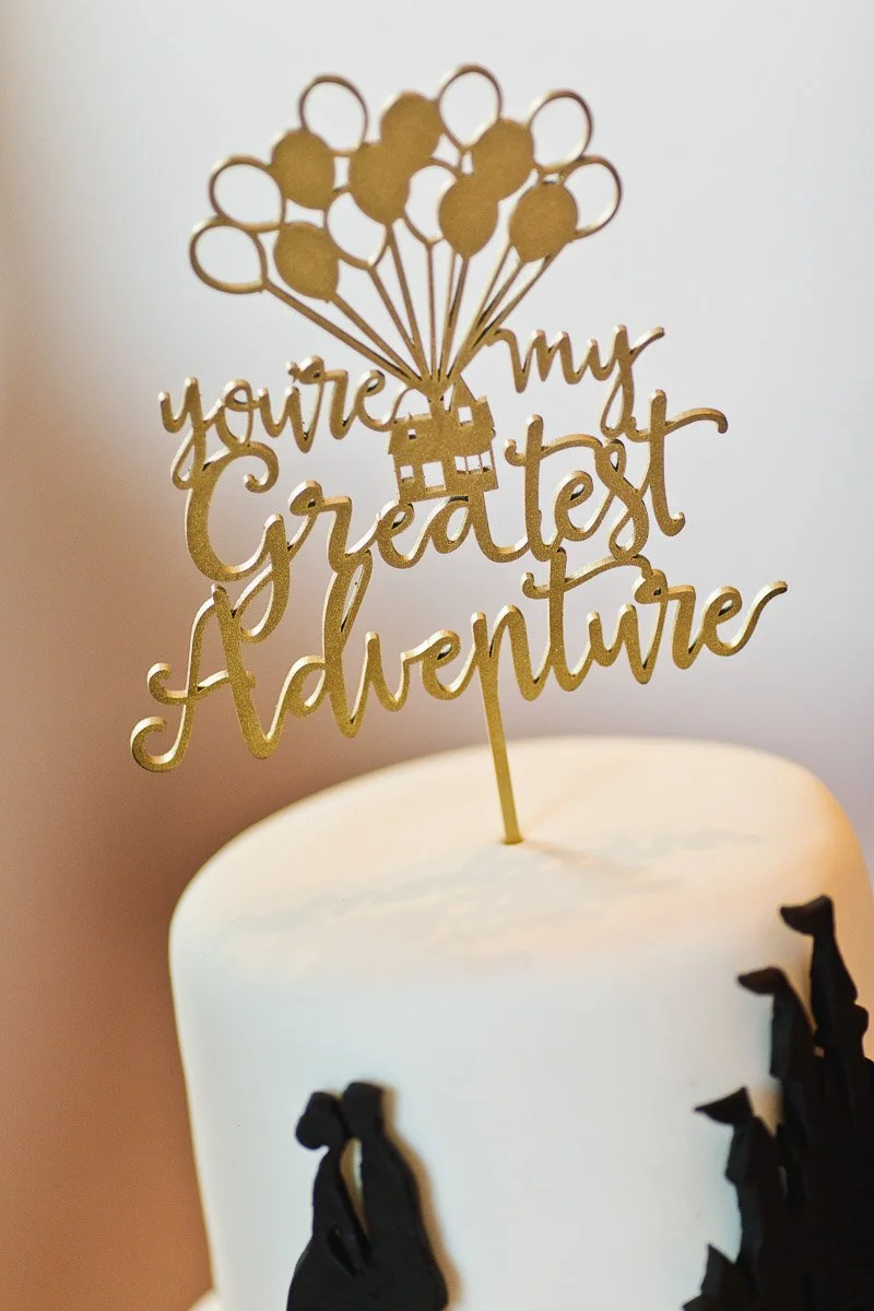 Wedding cake with a gold topper saying "you're my greatest adventure" and silhouetted figures on the white icing, evoking romance and discovery.