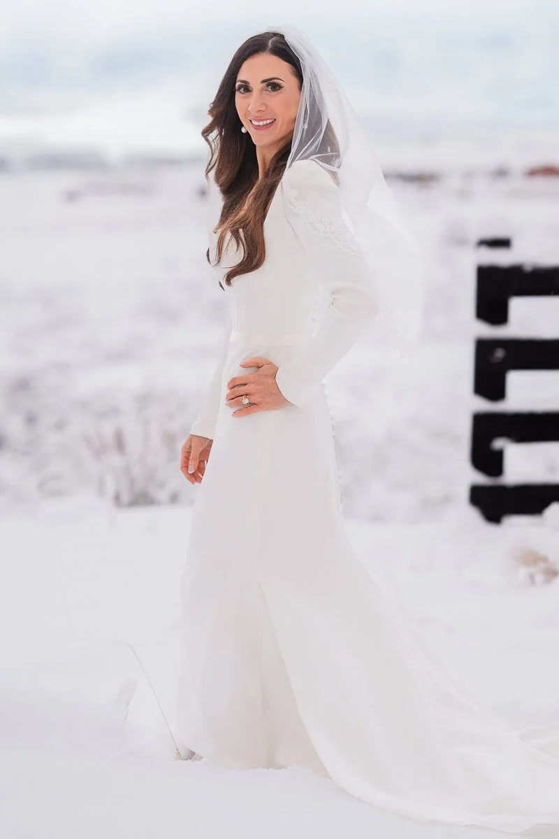 A smiling bride in a flowing white dress and veil stands in snow, with softly blurred mountains in the background, creating a serene and joyful scene.