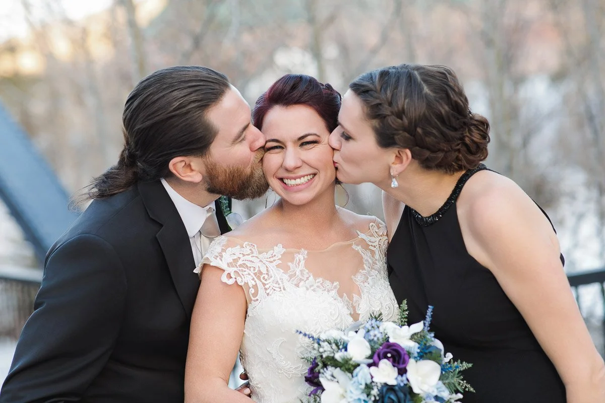 A bride in a lace wedding dress smiles brightly as she receives kisses on both cheeks from a bearded man in a suit and a woman in a black dress holding a bouquet.