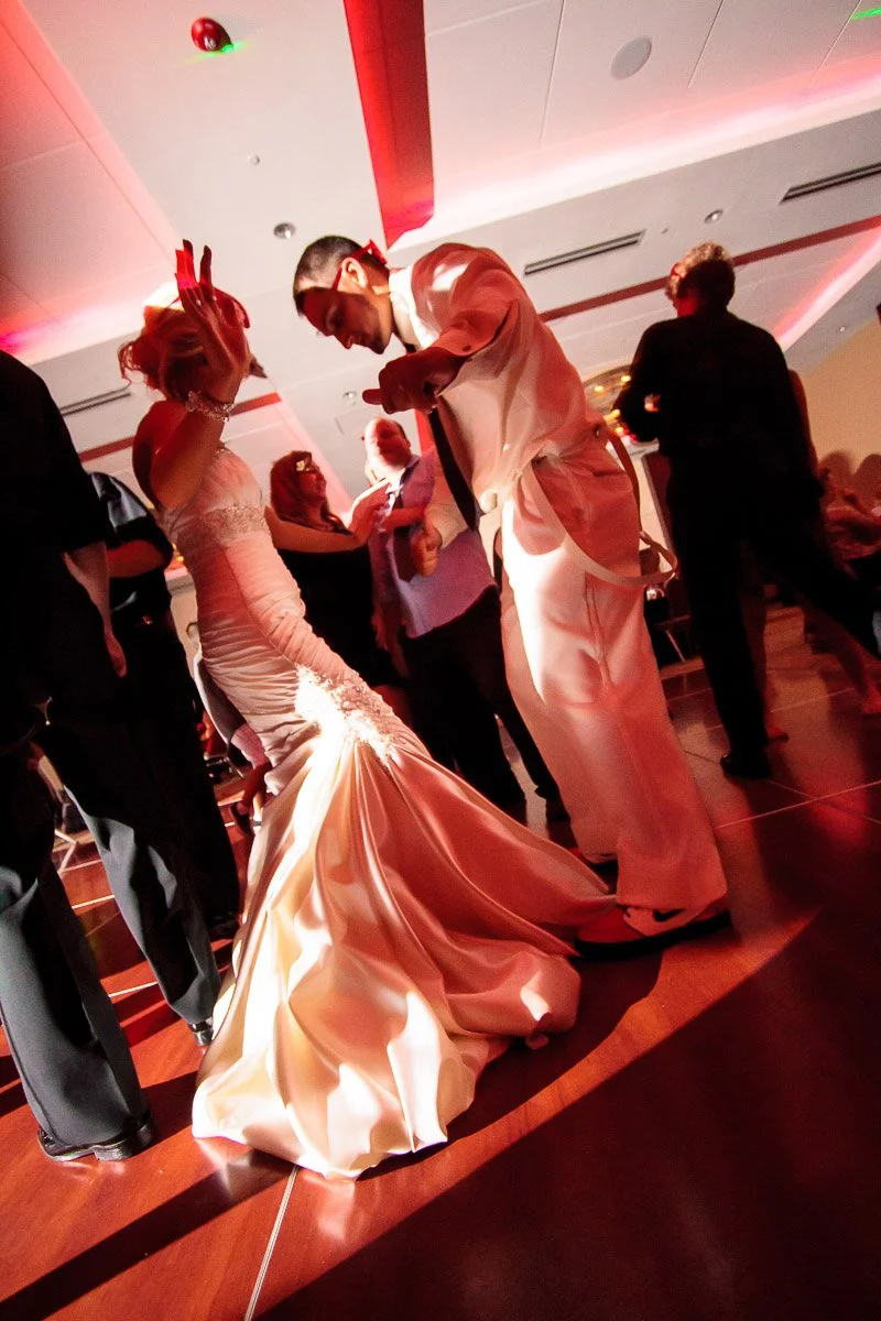 A bride and groom joyfully dance at a wedding reception. The bride's elegant gown contrasts with the groom's white suit. Warm lighting creates a festive mood.
