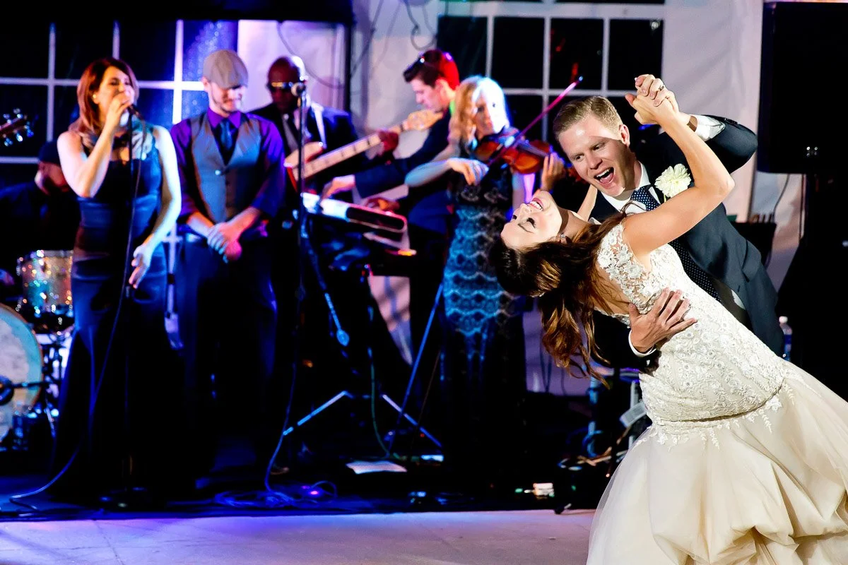 A bride and groom joyfully dance under the tent at a Keystone Ranch wedding while a live band performs in the background. The bride is in a white gown, and colorful stage lights create a lively atmosphere.