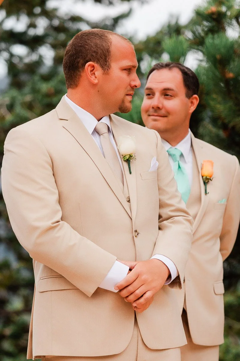 Two men in beige suits stand in front of trees. The front man gazes sideways with a serious expression, while the other smiles warmly at him.
