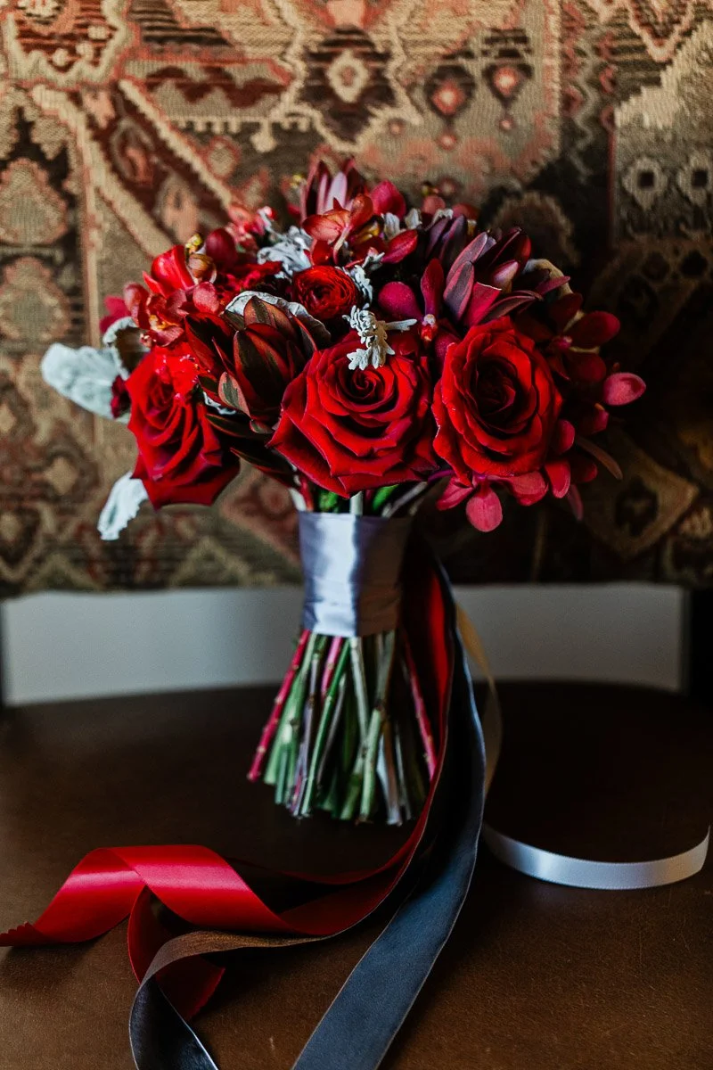A vibrant bouquet of red roses and flowers with green stems, wrapped in a white ribbon with red and dark ribbons. Background is a woven tapestry.