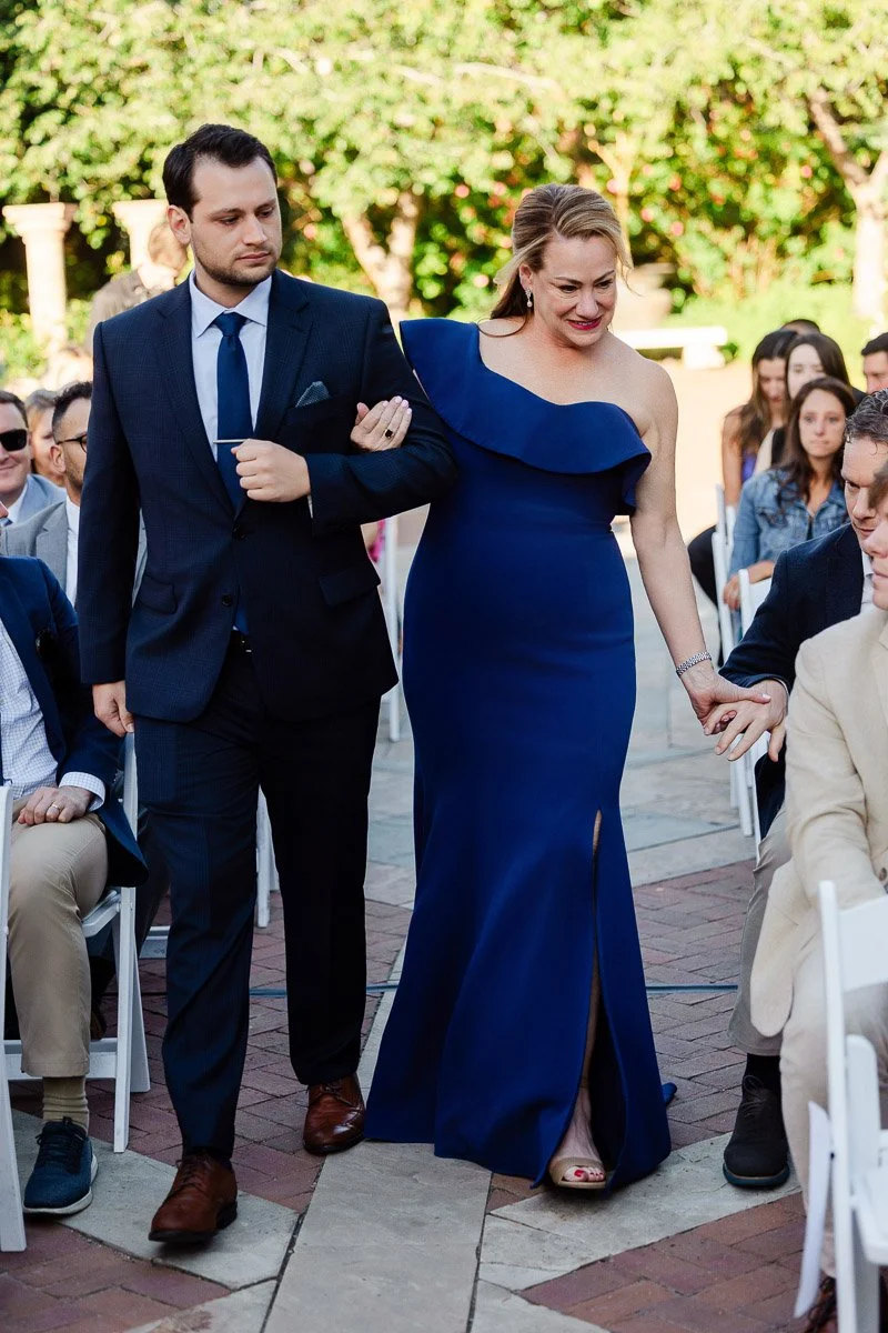 A man in a dark suit escorts a smiling woman in a blue, one-shoulder gown down a brick path at an outdoor event, surrounded by seated guests.