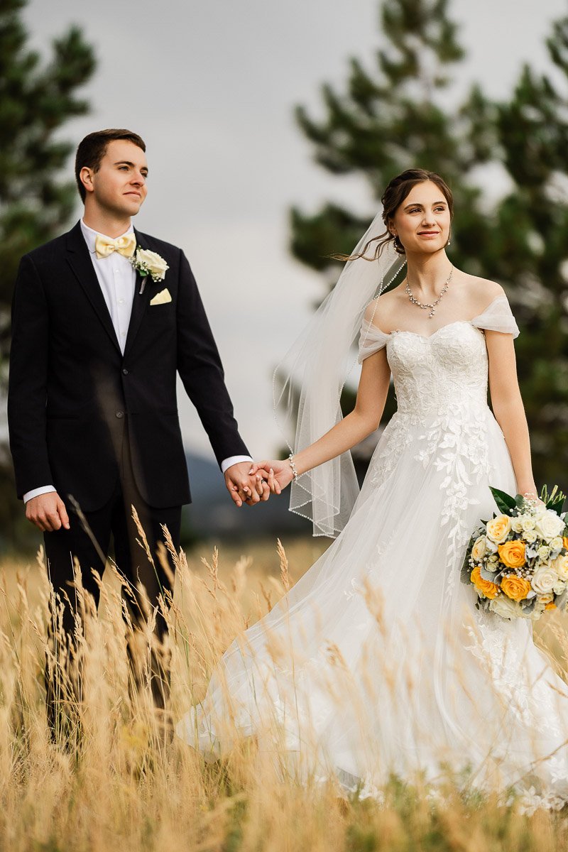 A bride and groom stand in a field of tall grass, holding hands. The bride wears a lace gown and veil, holding yellow roses. The groom is in a black suit.