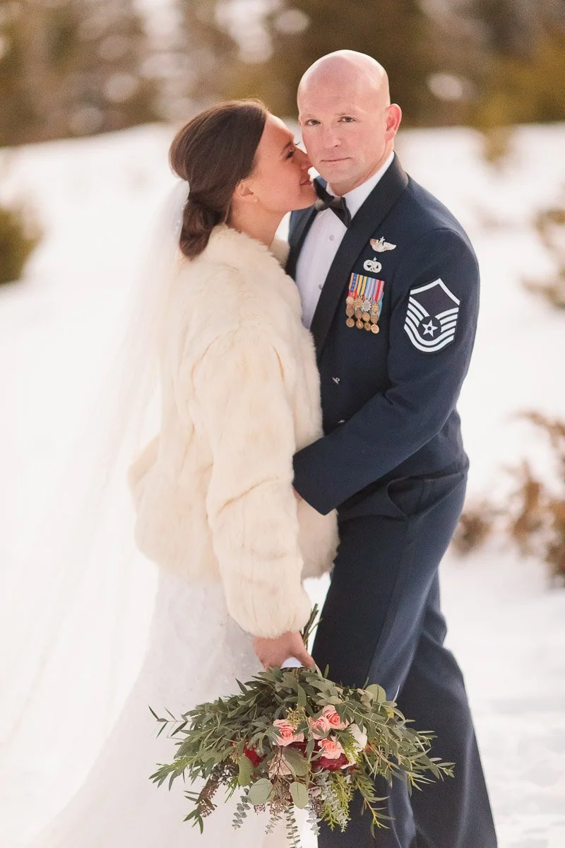 A bride in a white dress and fur shawl kisses a groom in a military uniform. They stand in a snowy landscape, holding a bouquet of pink flowers.