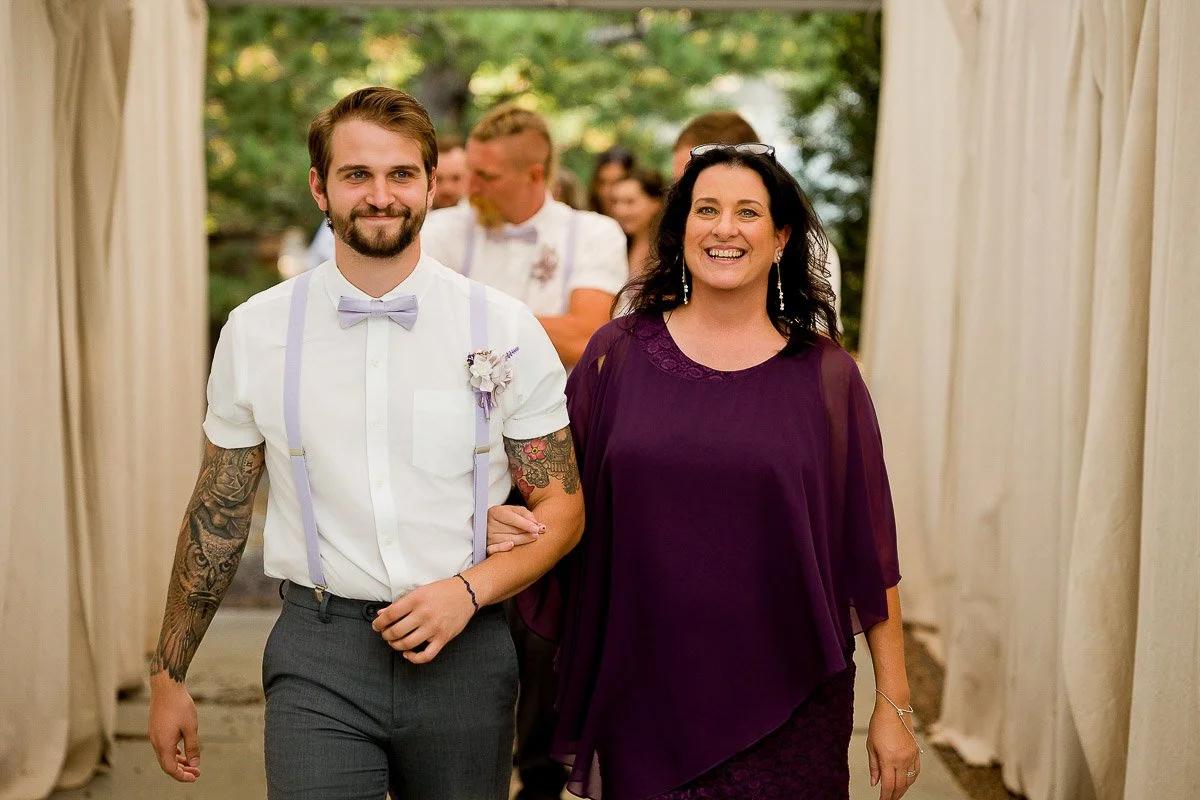 A smiling man in a white shirt with tattoos and a bow tie walks arm-in-arm with a woman in a purple dress. They appear joyful, against a blurred outdoor background.