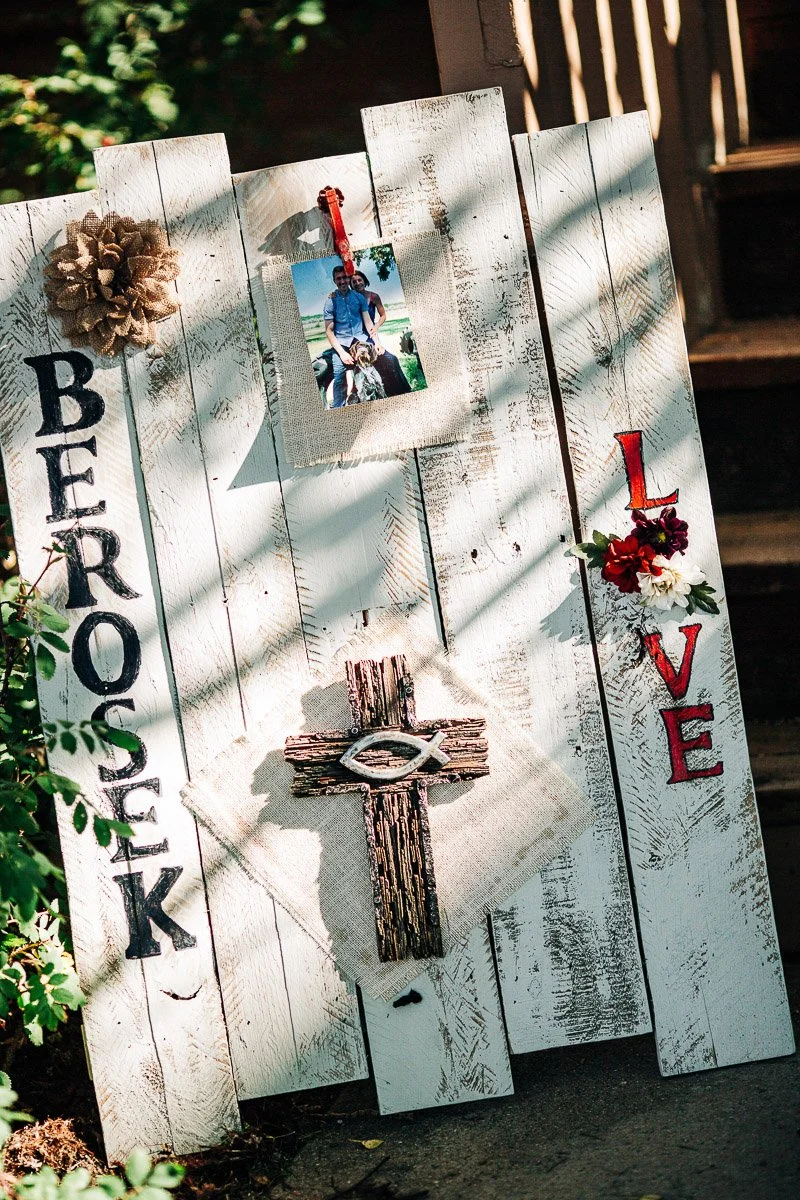A rustic white wooden sign with "Berosek" and "Love" written in bold letters. A photo of a person with a dog is clipped above a cross with a fish symbol. Sunlight casts dappled shadows, creating a serene, reflective atmosphere. Flowers adorn the sign