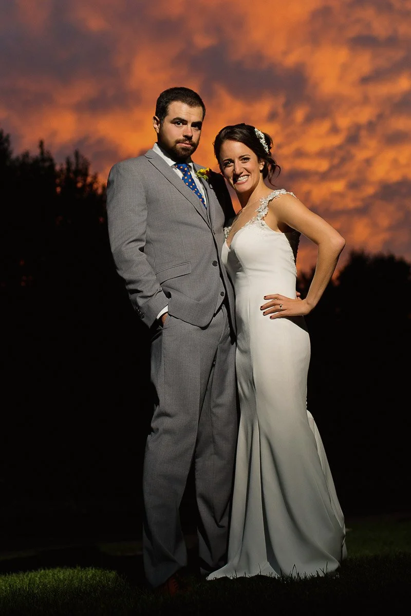 A bride and groom pose outdoors during sunset. The bride wears a white gown, the groom a gray suit. The sky is vividly orange, creating a dramatic backdrop.