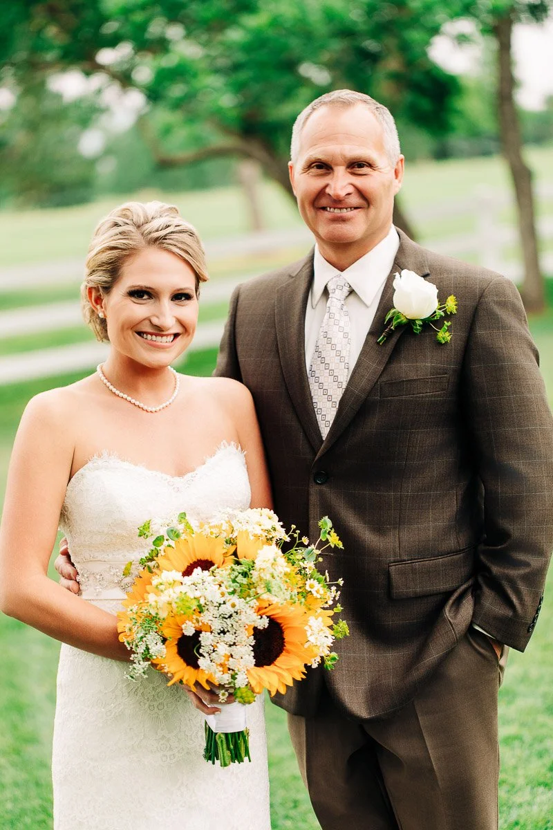 

A smiling bride in a white lace dress holds a sunflower bouquet beside a man in a brown suit with a white flower boutonniere. Greenery in the background.