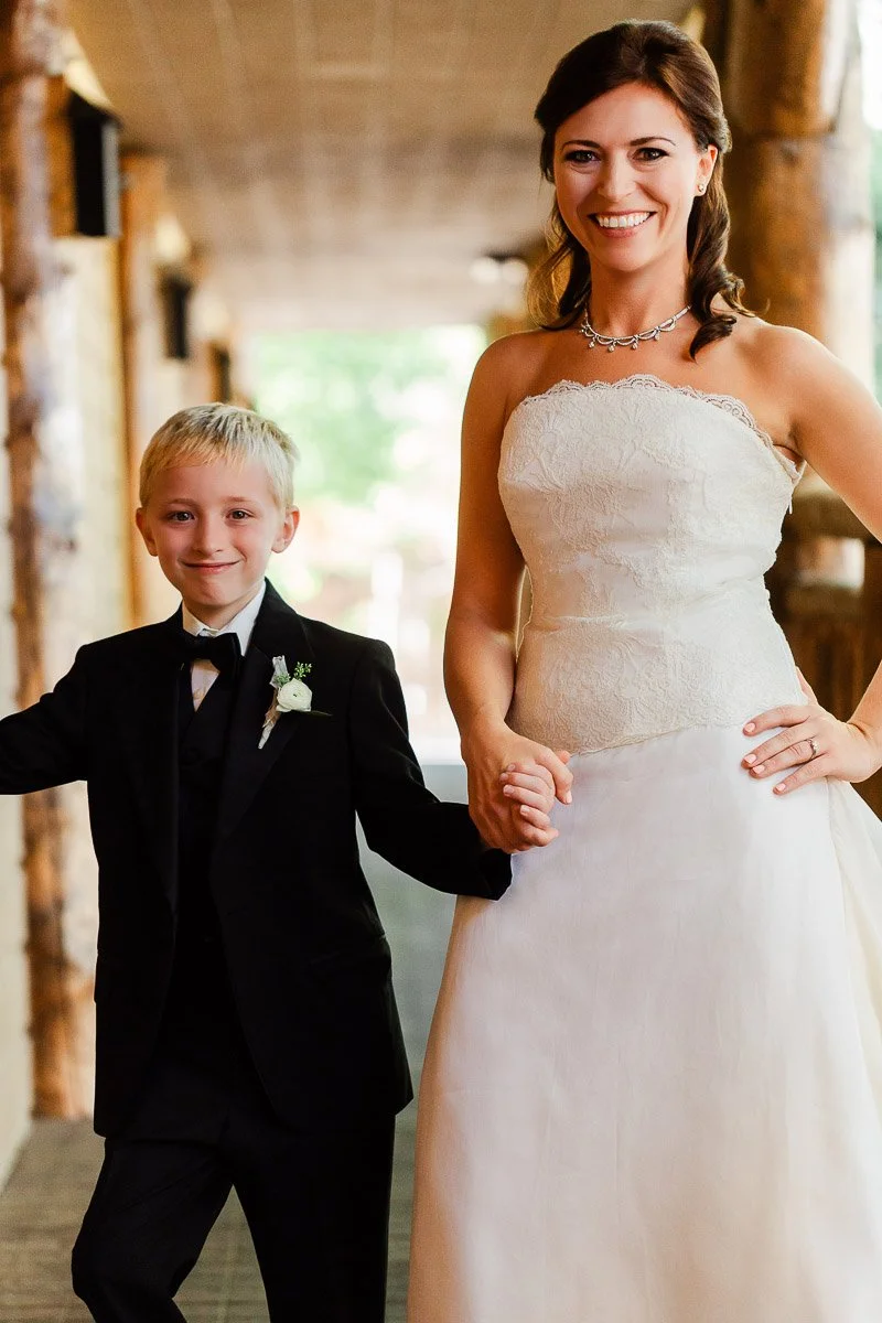 A smiling woman in a white strapless wedding dress holds hands with a young boy in a black suit and bow tie. They stand in a warmly lit hallway.