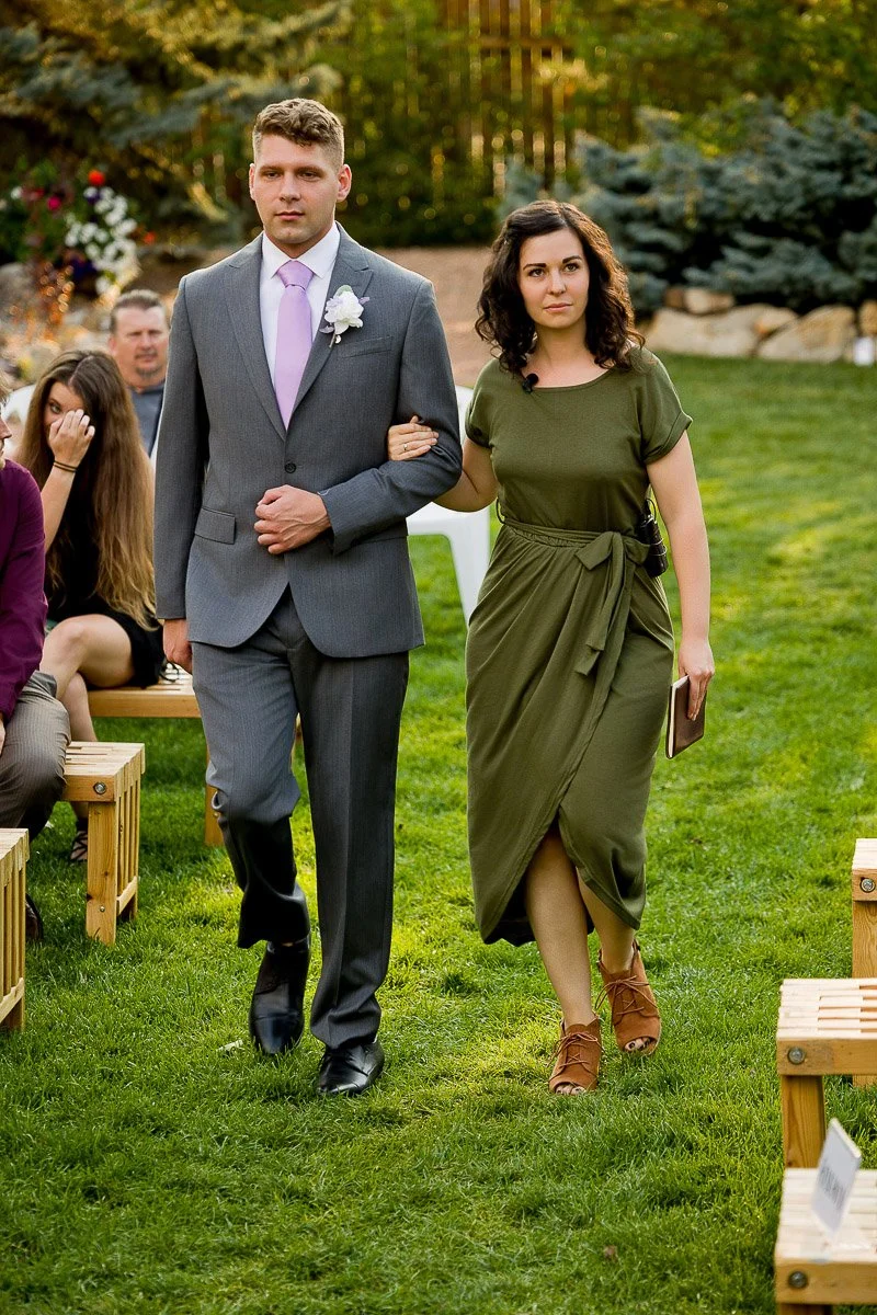A man in a gray suit with a pink tie and white boutonniere walks arm in arm with a woman in a green dress holding a book. They are outdoors on green grass with benches and a backdrop of trees and flowers, conveying a formal and serene atmosphere.