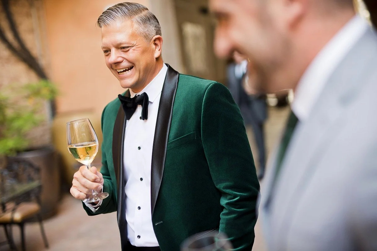 Man in a green velvet tuxedo and bow tie holds a wine glass, smiling warmly. The scene is festive, with another person in a gray suit nearby.
