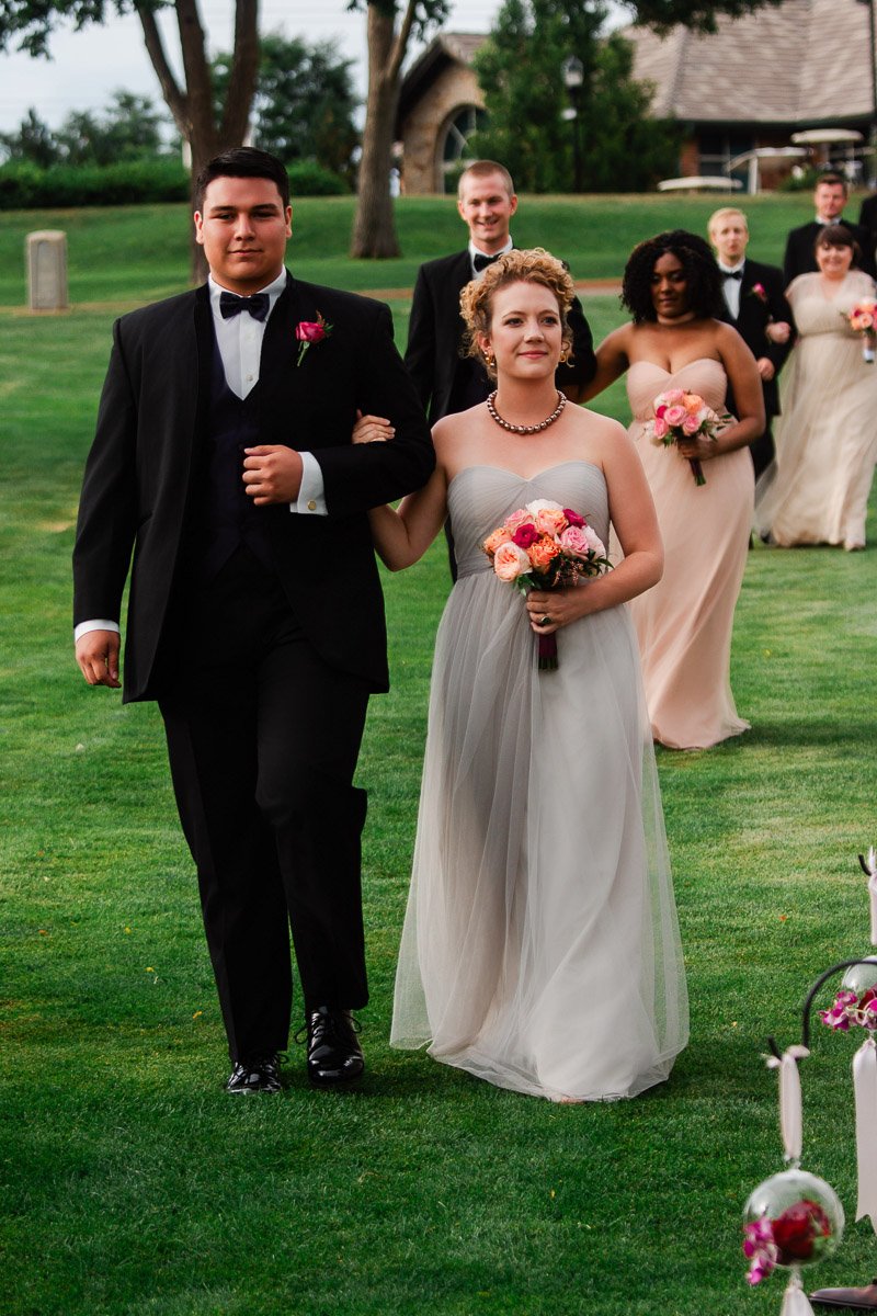 A wedding party walks across a lush green lawn. The lead couple is elegantly dressed; the woman holds a bouquet of pink and orange flowers, evoking joy.