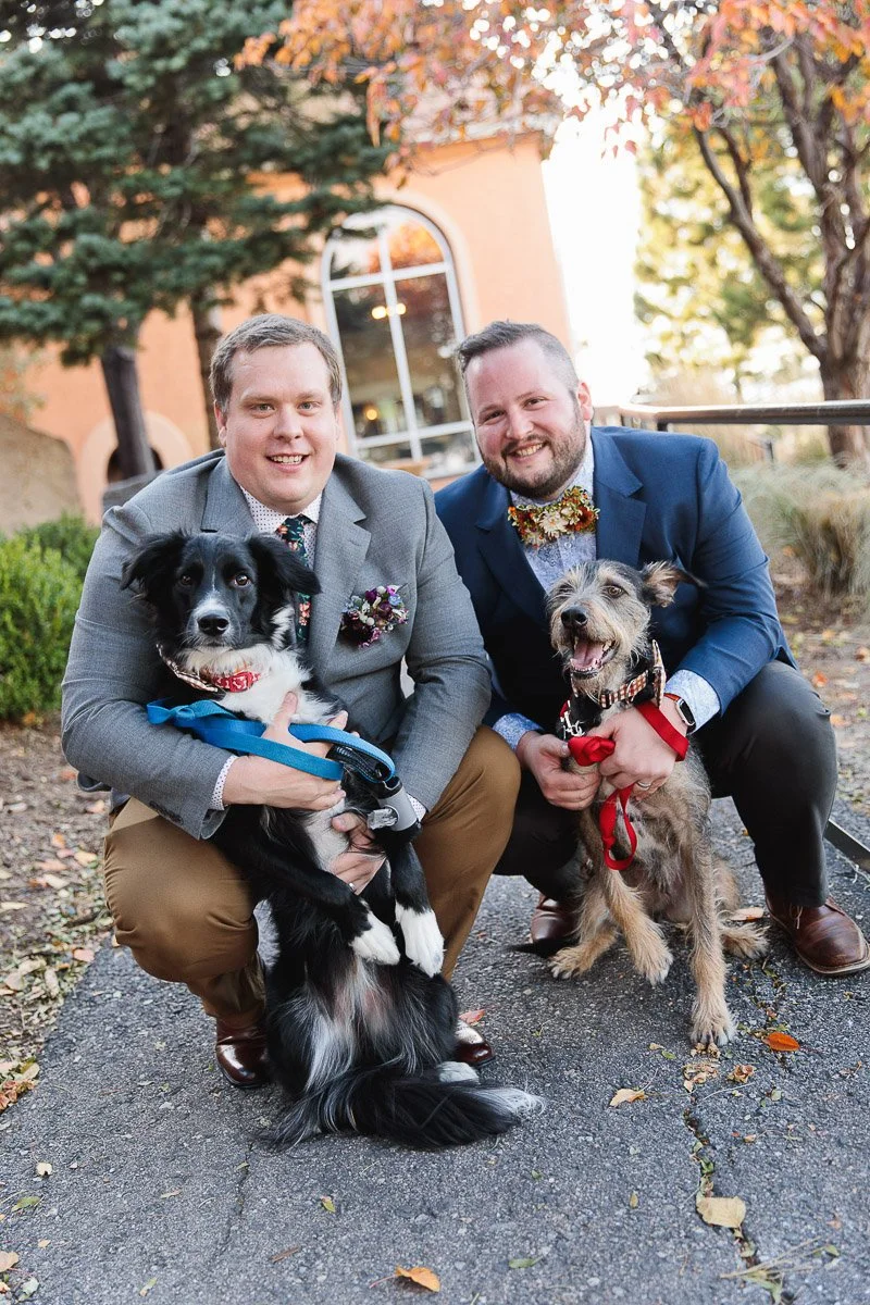 Two men in suits kneel beside each other outdoors, each holding a dog. They smile warmly under autumn trees, creating a joyful, celebratory scene.