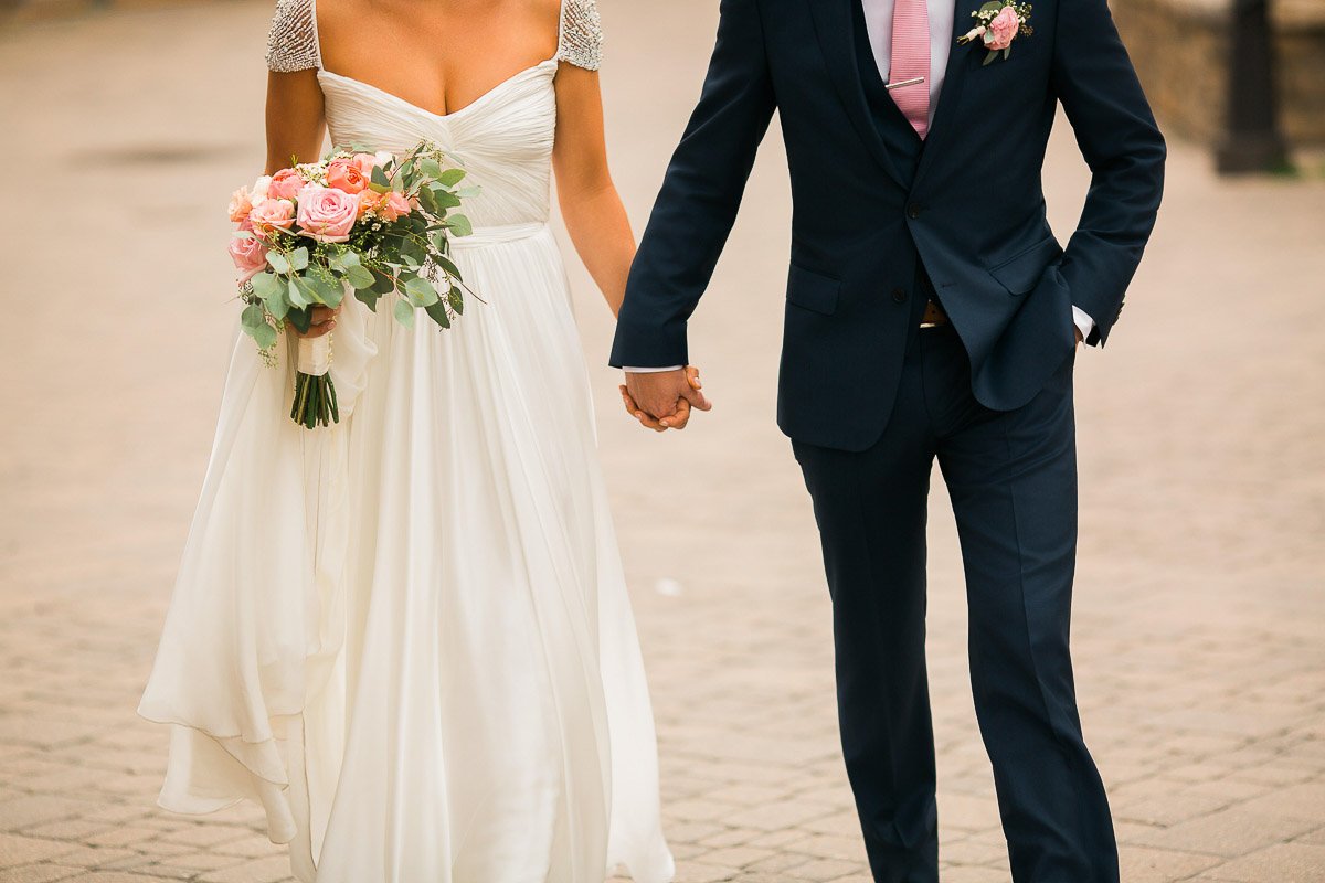 Bride and groom holding hands, walking on a cobblestone path. Bride in a white dress with pink bouquet, groom in navy suit with pink tie. Celebratory mood.
