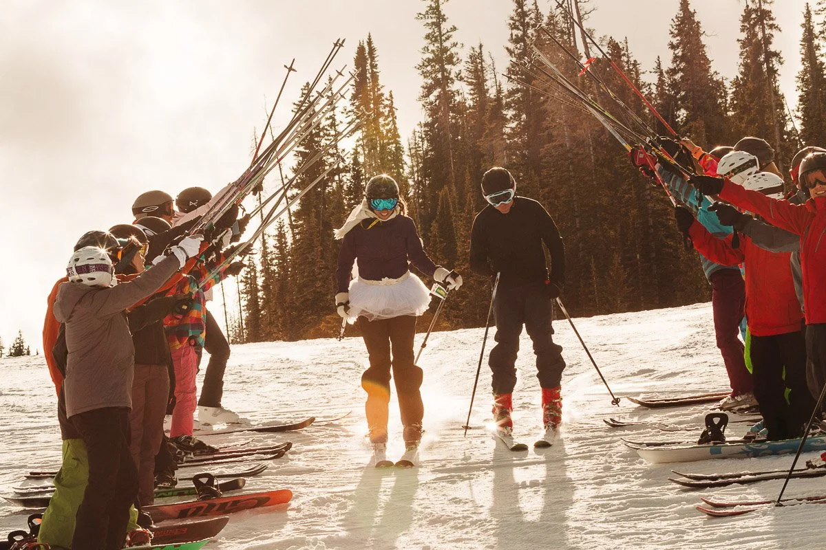 A group of skiers in colorful outfits form an arch with their poles on a snowy slope. Two skiers, one wearing a tutu, joyfully ski through. The scene is bright and cheerful, with tall pine trees in the background.
