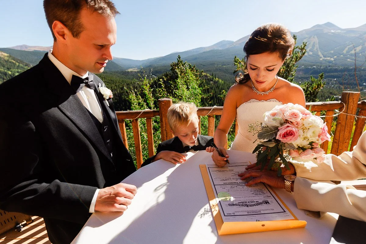Bride and groom in formal attire sign a document outdoors with a child observing. The bride holds a bouquet. Mountain view in the background.