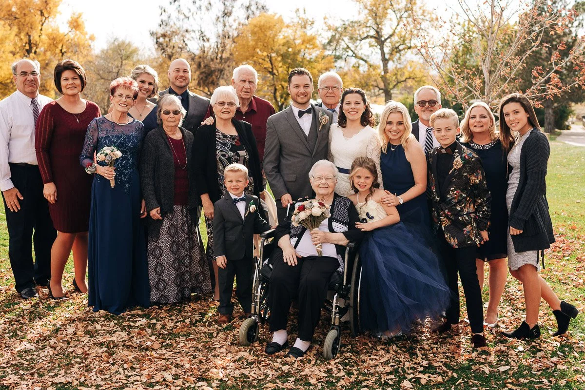 A joyful family gathers outdoors for a group photo on a bright autumn day. The setting is warm, with fallen leaves and vibrant yellow trees in the background. Front and center is an elderly woman in a wheelchair, holding a bouquet. A man in a suit an