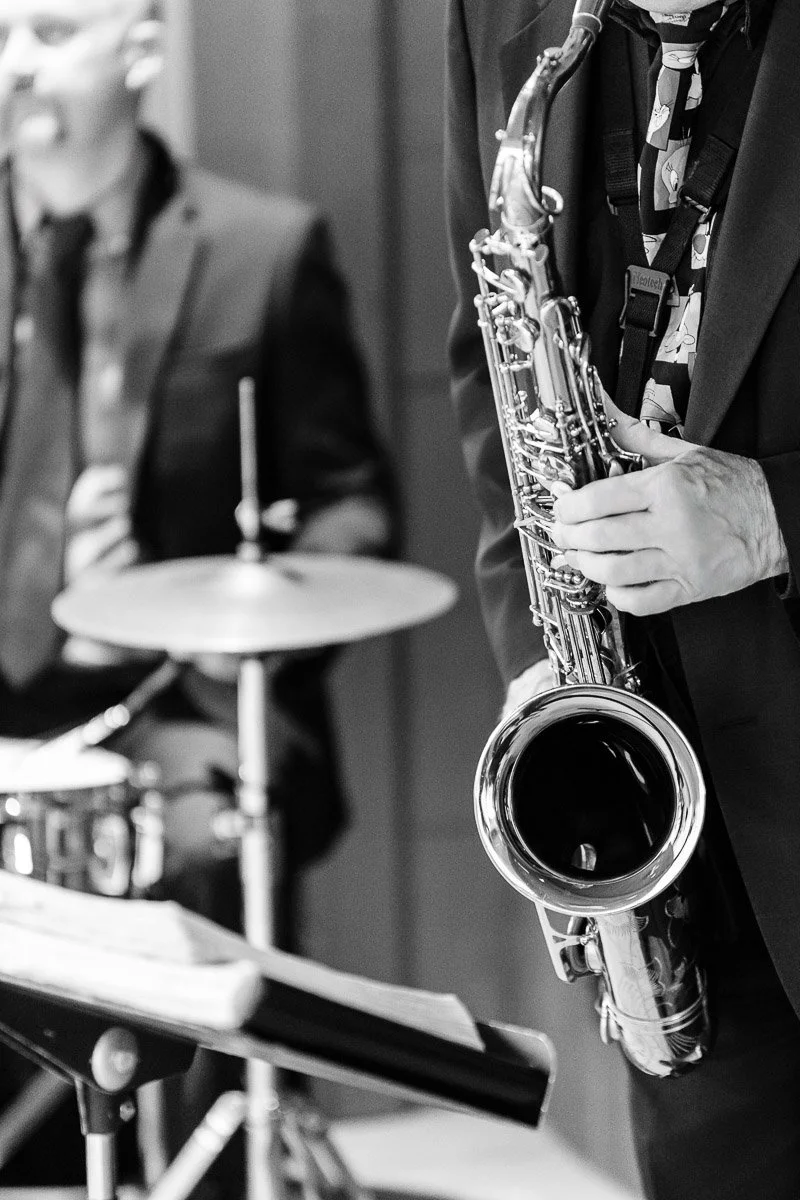 Black and white image of a jazz musician playing a saxophone, alongside a drummer. Both are in suits, creating a classic, elegant atmosphere.