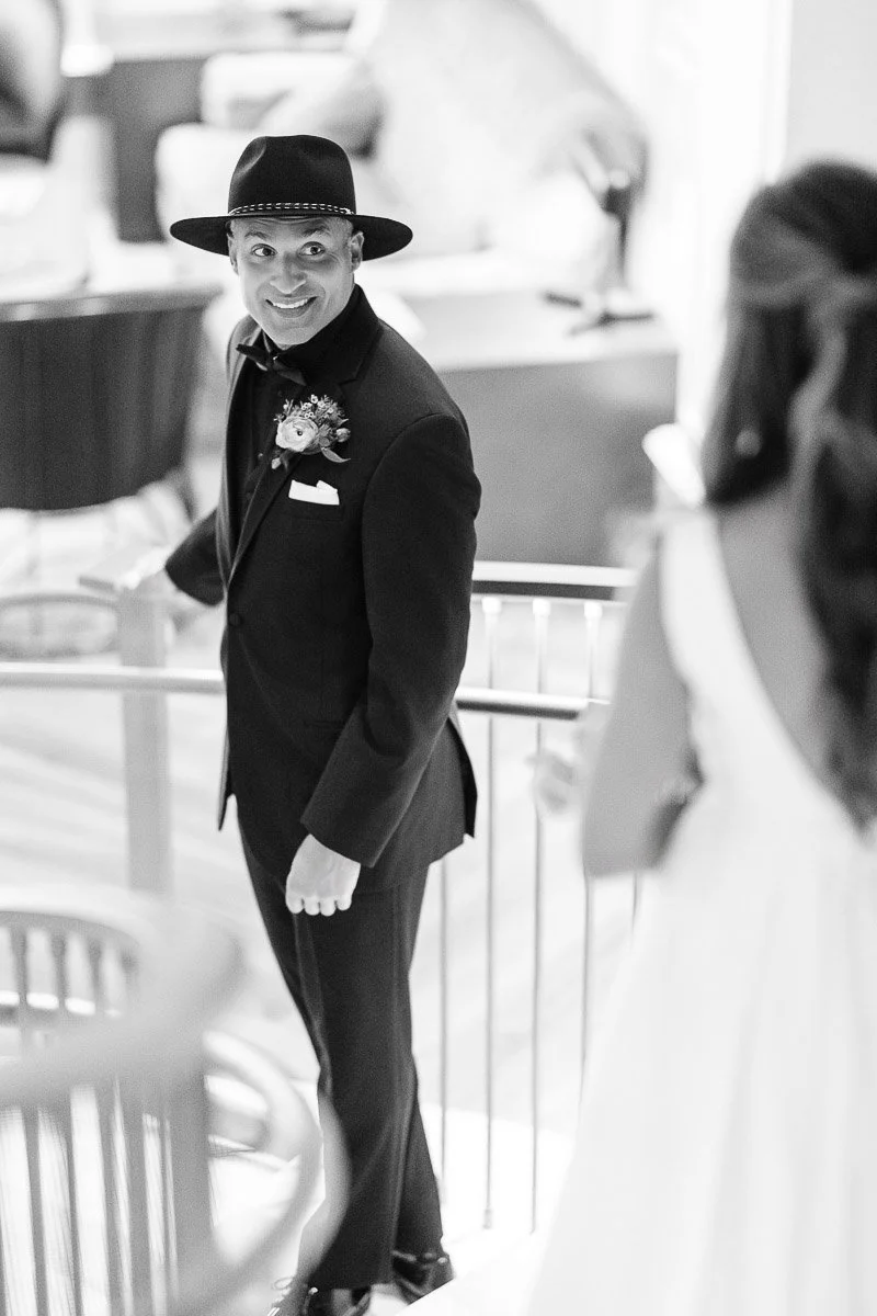 A groom in a suit and hat smiles joyfully as he sees the bride, who is dressed in white, ascending a staircase. The mood is warm and celebratory.