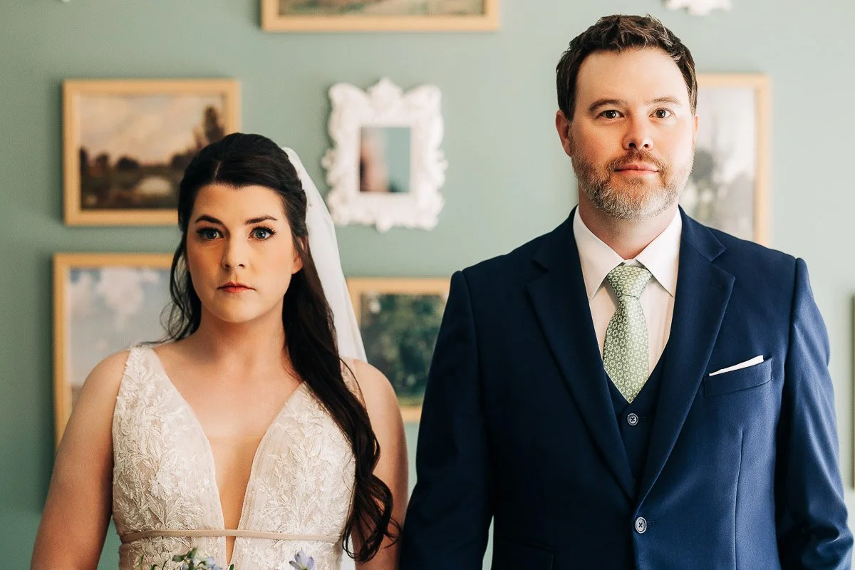 A bride and groom stand side by side, facing forward. The bride wears a lacy white dress and veil, while the groom is in a navy suit with a green tie. They look serious, with framed art on a green wall behind them.