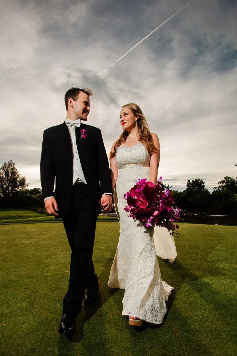 A bride in a white lace gown and groom in a black tuxedo walk hand in hand on grass, under a cloudy sky. She holds a bouquet of vibrant purple flowers.