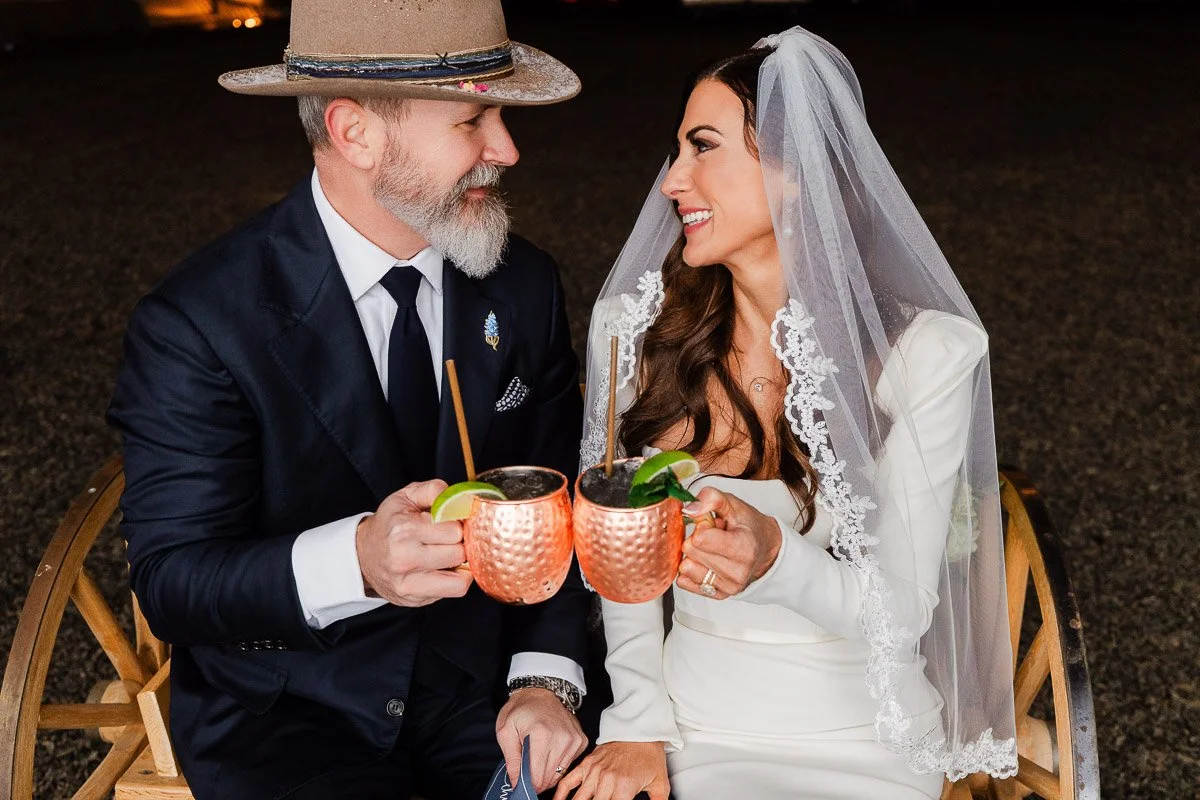 A bride in a white dress and veil and a groom in a suit with a hat toast with copper mugs, smiling warmly at each other. Romantic and joyful scene captured by La Joya Dulce wedding photographer tomKphoto
