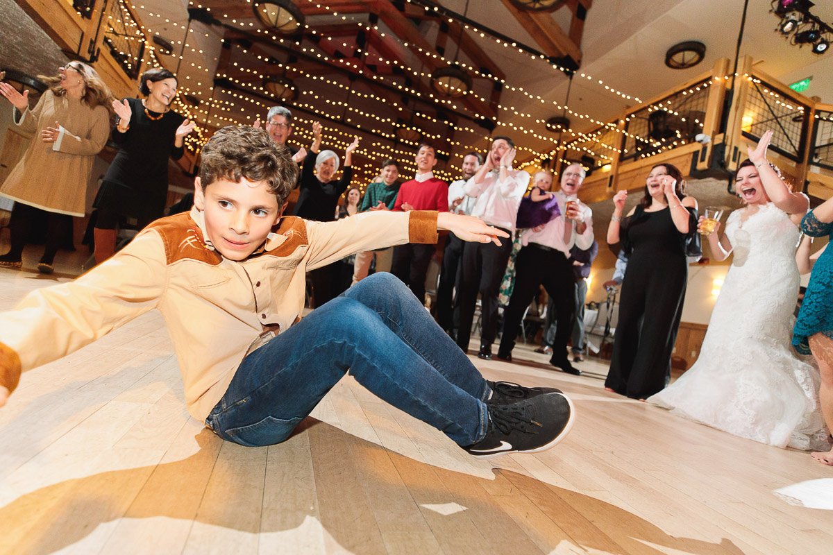 A young boy in a beige and brown shirt breakdances energetically on a wooden floor at a festive event. A cheering crowd, including a bride, surrounds him under warm string lights, creating an atmosphere of celebration and joy.