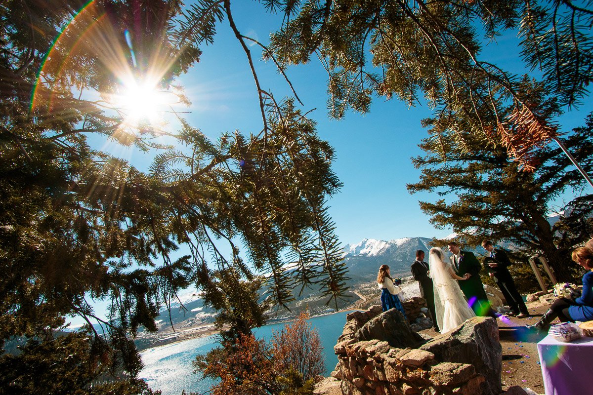 Wedding ceremony under a bright sun, framed by tree branches. A couple stands with the officiant near a scenic lake and mountain vista, creating a romantic atmosphere.