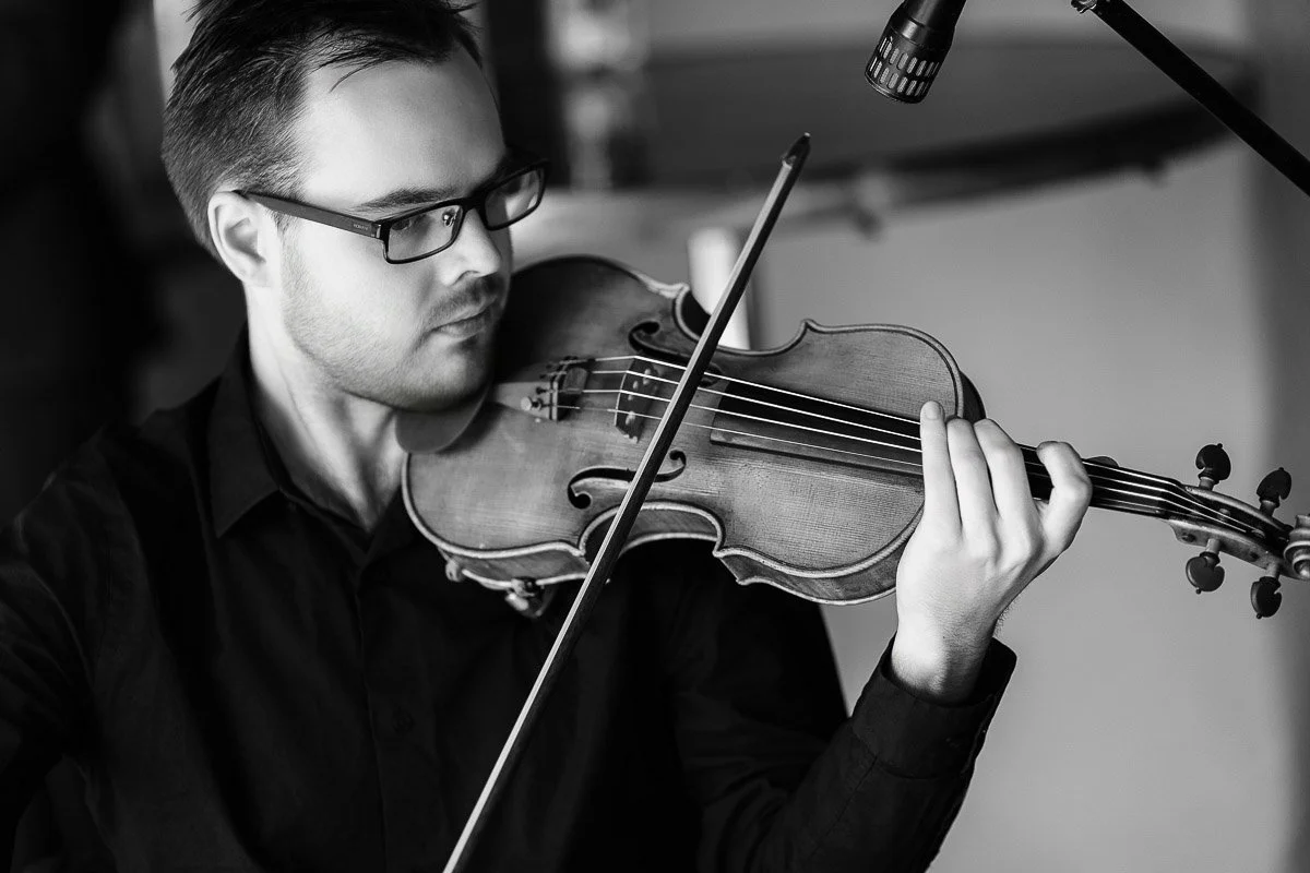 A focused musician in glasses plays the violin, wearing a dark shirt. The black-and-white photo conveys a serene and passionate ambiance.